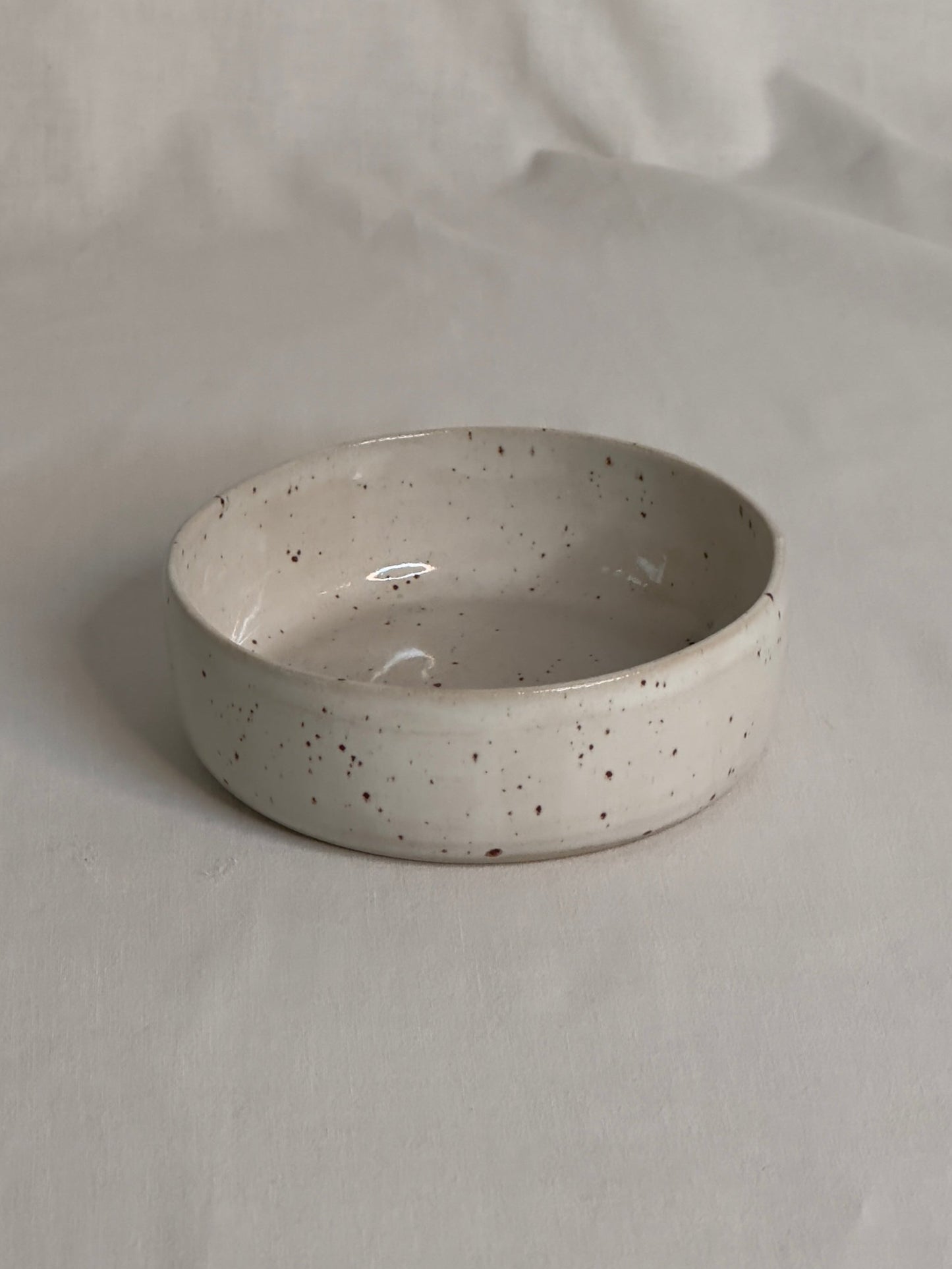 Bol en céramique artisanale blanc moucheté, avec un charme rustique. Sa forme est ronde, avec des bords épais et légèrement arrondis. Le bol repose sur une surface de couleur claire, créant une atmosphère douce et minimaliste.