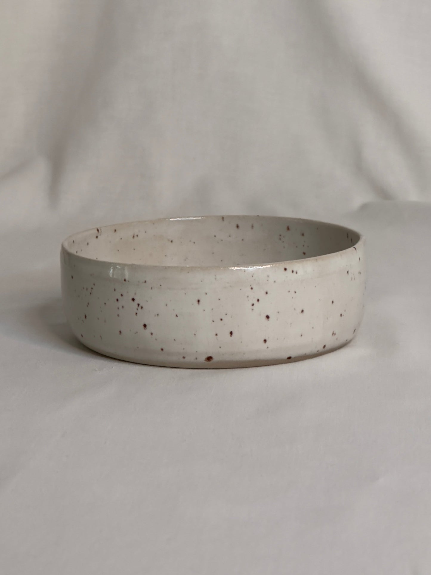 Bol en céramique artisanale blanc moucheté, avec un charme rustique. Sa forme est ronde, avec des bords épais et légèrement arrondis. Le bol repose sur une surface de couleur claire, créant une atmosphère douce et minimaliste.
