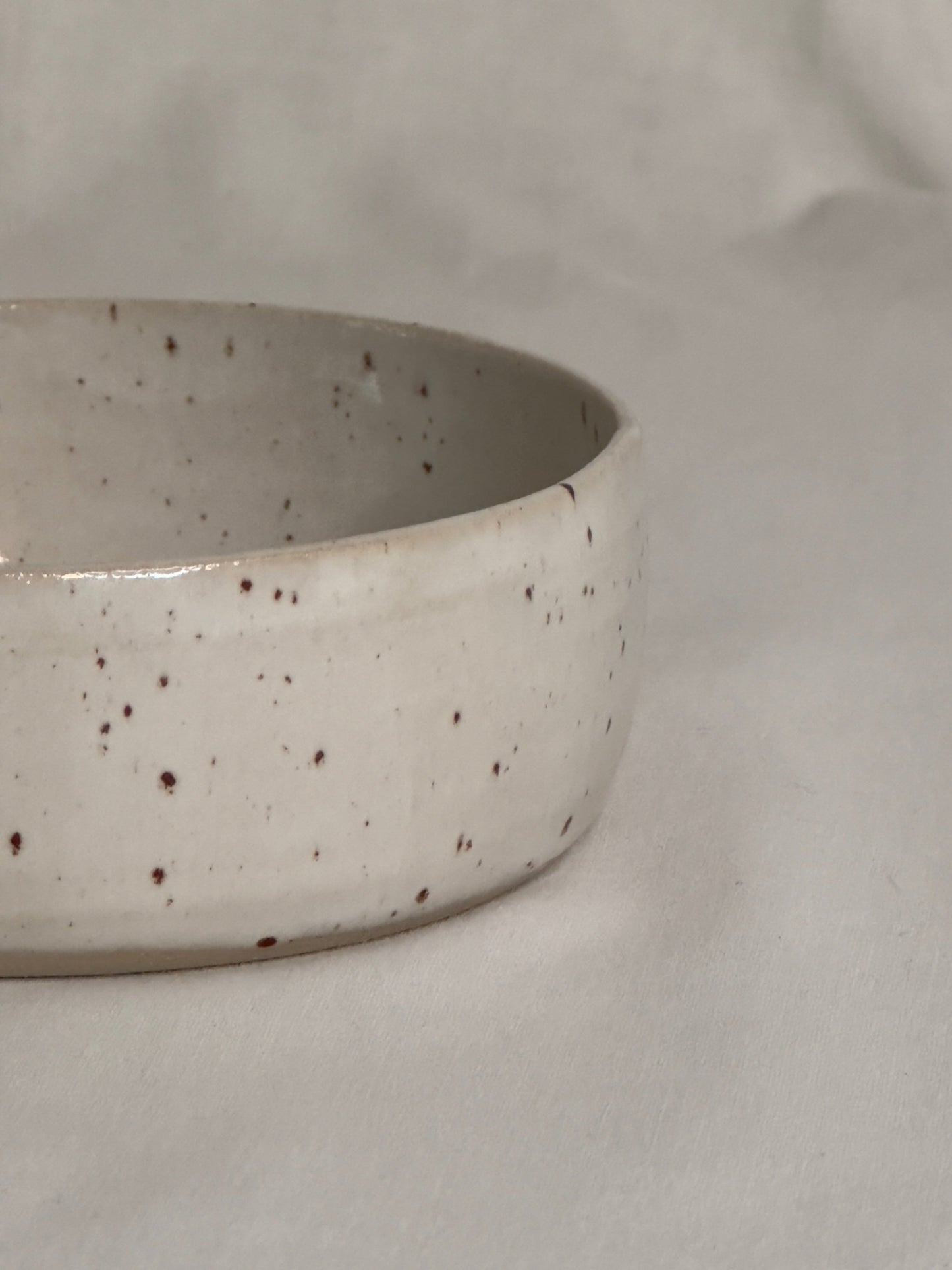 Bol en céramique artisanale blanc moucheté, avec un charme rustique. Sa forme est ronde, avec des bords épais et légèrement arrondis. Le bol repose sur une surface de couleur claire, créant une atmosphère douce et minimaliste.