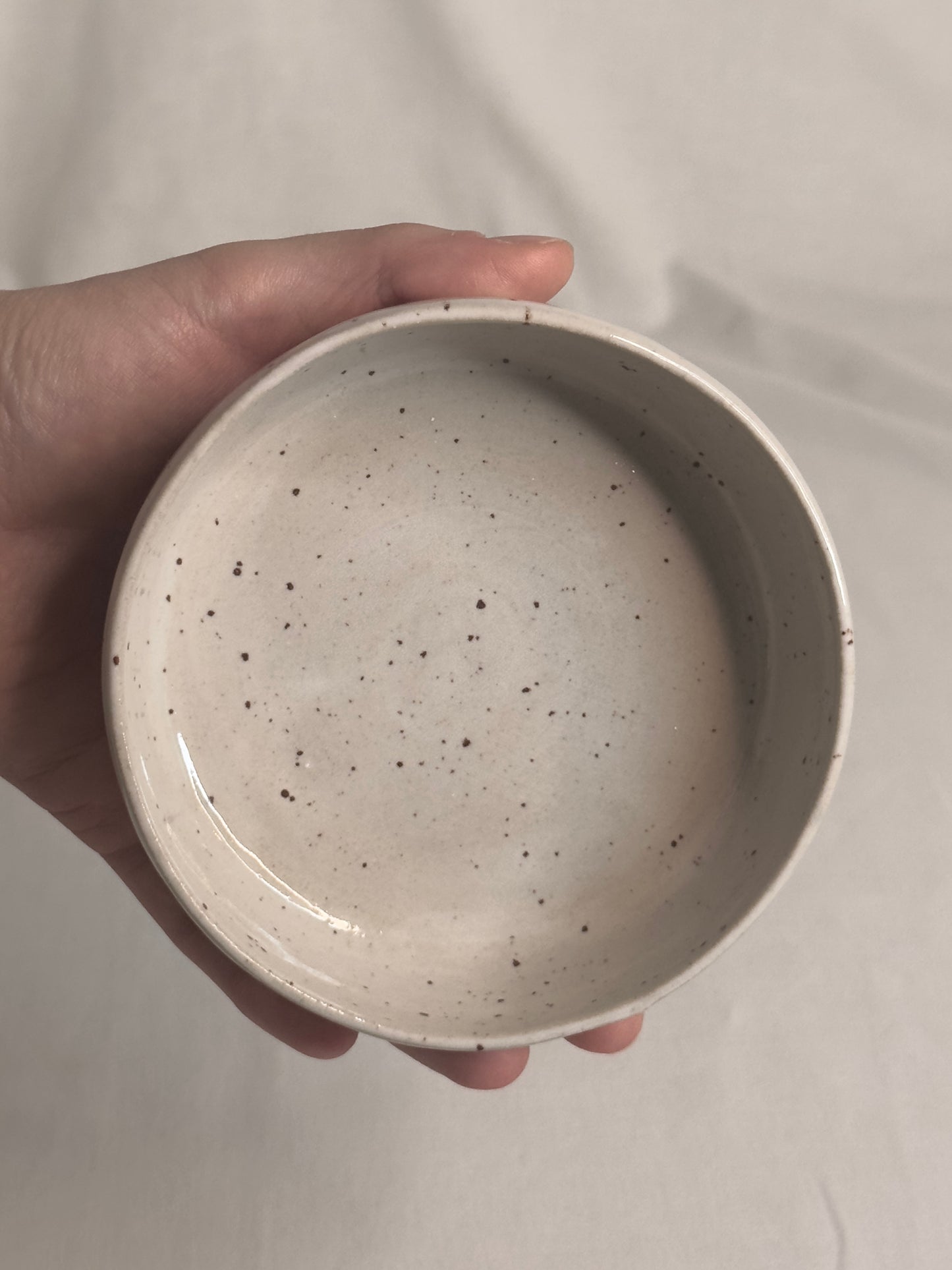 Bol en céramique artisanale blanc moucheté, avec un charme rustique. Sa forme est ronde, avec des bords épais et légèrement arrondis. Le bol repose sur une surface de couleur claire, créant une atmosphère douce et minimaliste.