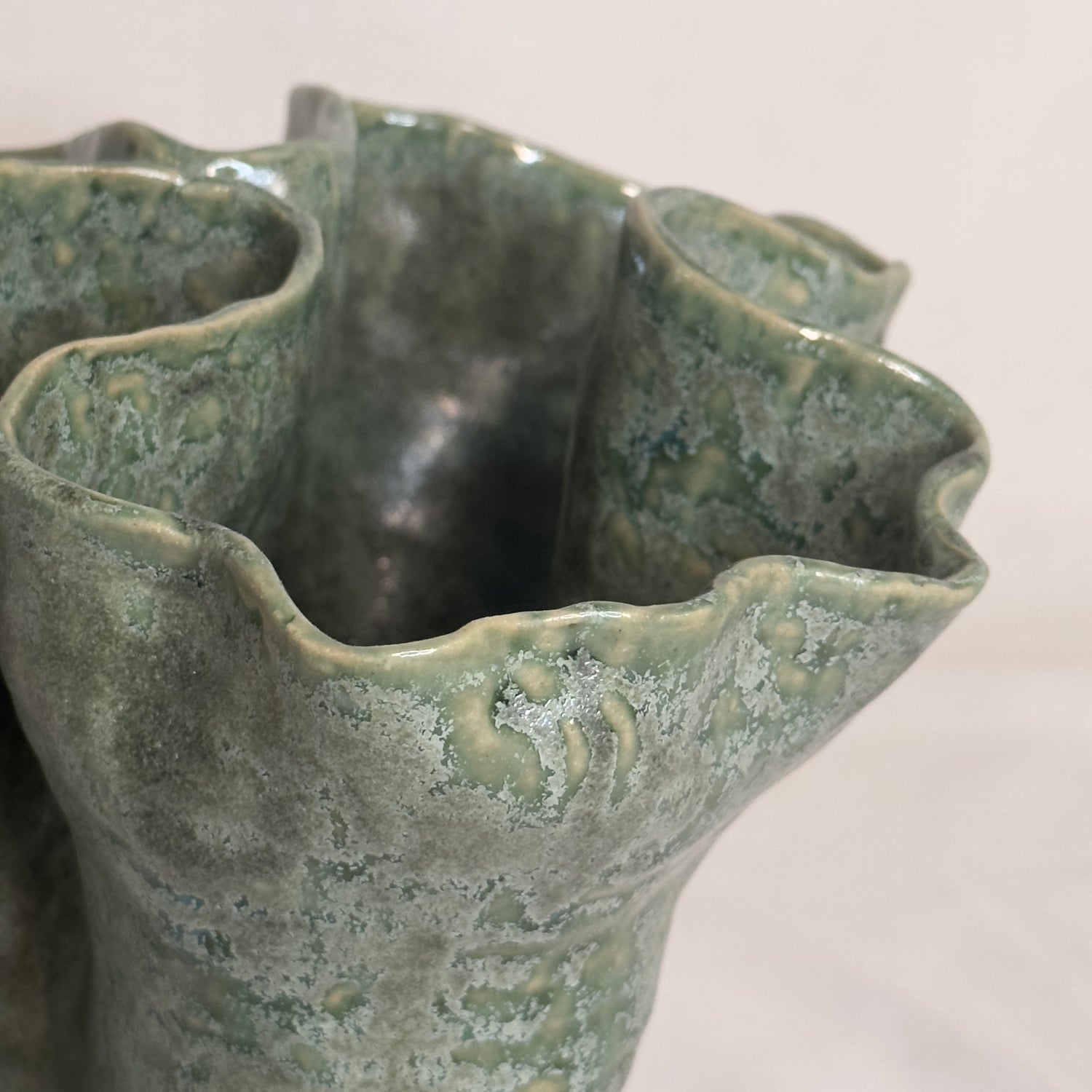 Vase en céramique sculpté à la main, présentant une forme organique et asymétrique. La surface texturée est recouverte d'une glaçure vert marbré avec des nuances de gris et des reflets brillants. L'ouverture du vase est large et irrégulière, soulignant son caractère artisanal et unique. Le design rappelle des formes naturelles, idéal comme pièce décorative ou vase fonctionnel.