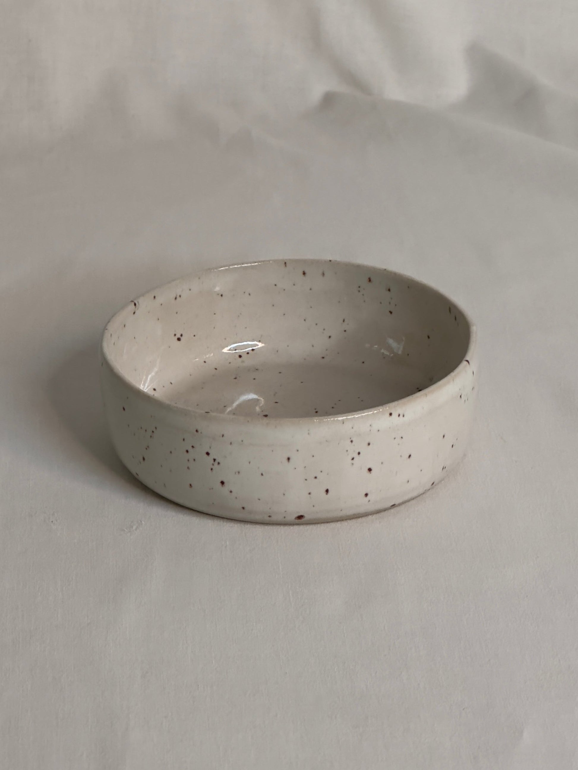 Bol en céramique artisanale blanc moucheté, avec un charme rustique. Sa forme est ronde, avec des bords épais et légèrement arrondis. Le bol repose sur une surface de couleur claire, créant une atmosphère douce et minimaliste.